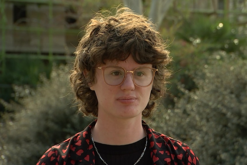 A person with short curly brown hair and glasses looks serious.