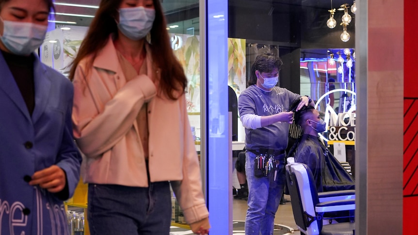 Un coiffeur coupe les cheveux d'un client alors que deux femmes masquées passent dans la rue