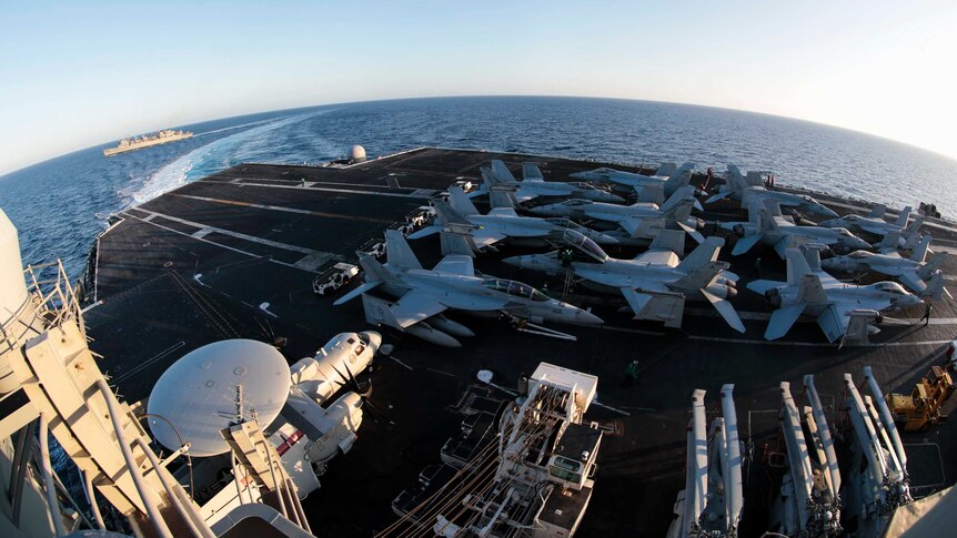 A wide shot sees fighter jets parked on an aircraft carrier's runway as another navy ship sails by in the background