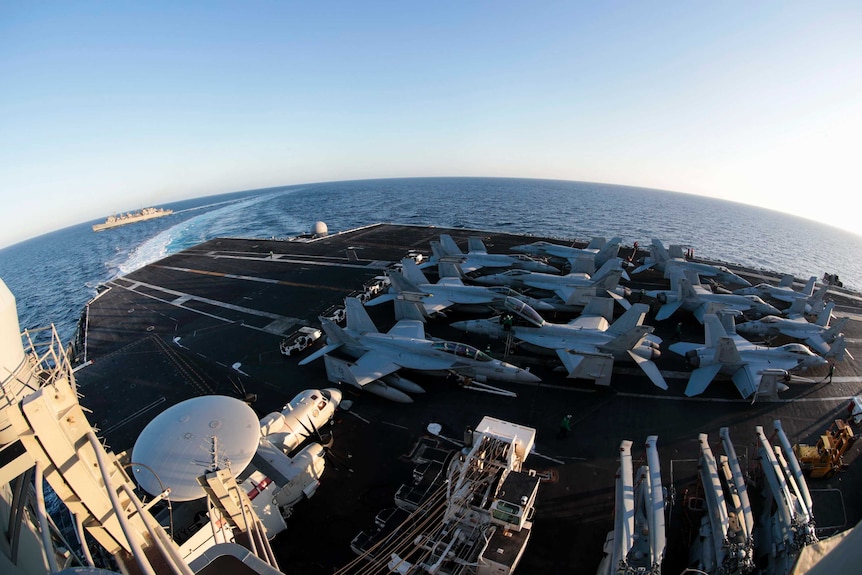 A wide shot sees fighter jets parked on an aircraft carrier's runway as another navy ship sails by in the background