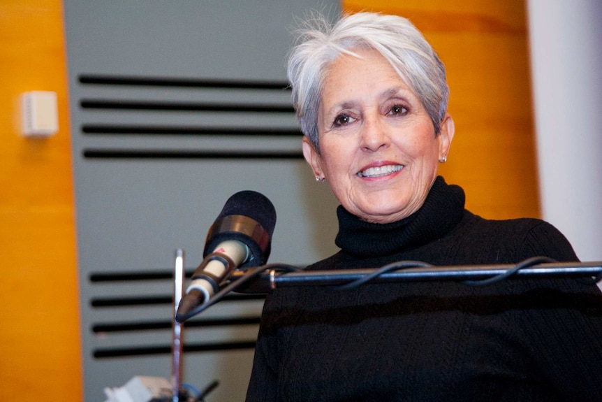 Singer Joan Baez smiling as she sits in front of the microphone looking just off camera.