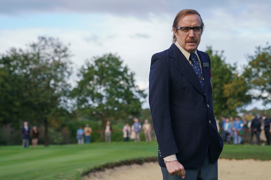 A blond middle-aged man, in a suit, tie and glasses, looks unimpressed, while standing on a golf course