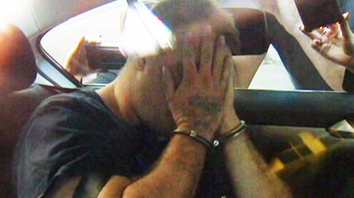 A handcuffed man covers his face with his hands as he sits in the back seat of a car.