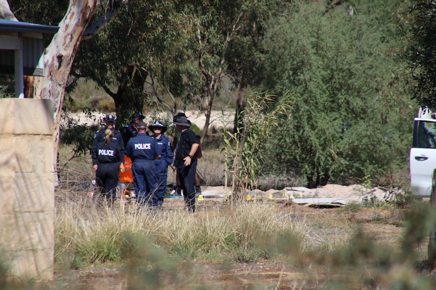 A wide shot showing a group of WA Police forensics officers huddled outdoors among shrubs and trees on a private property.