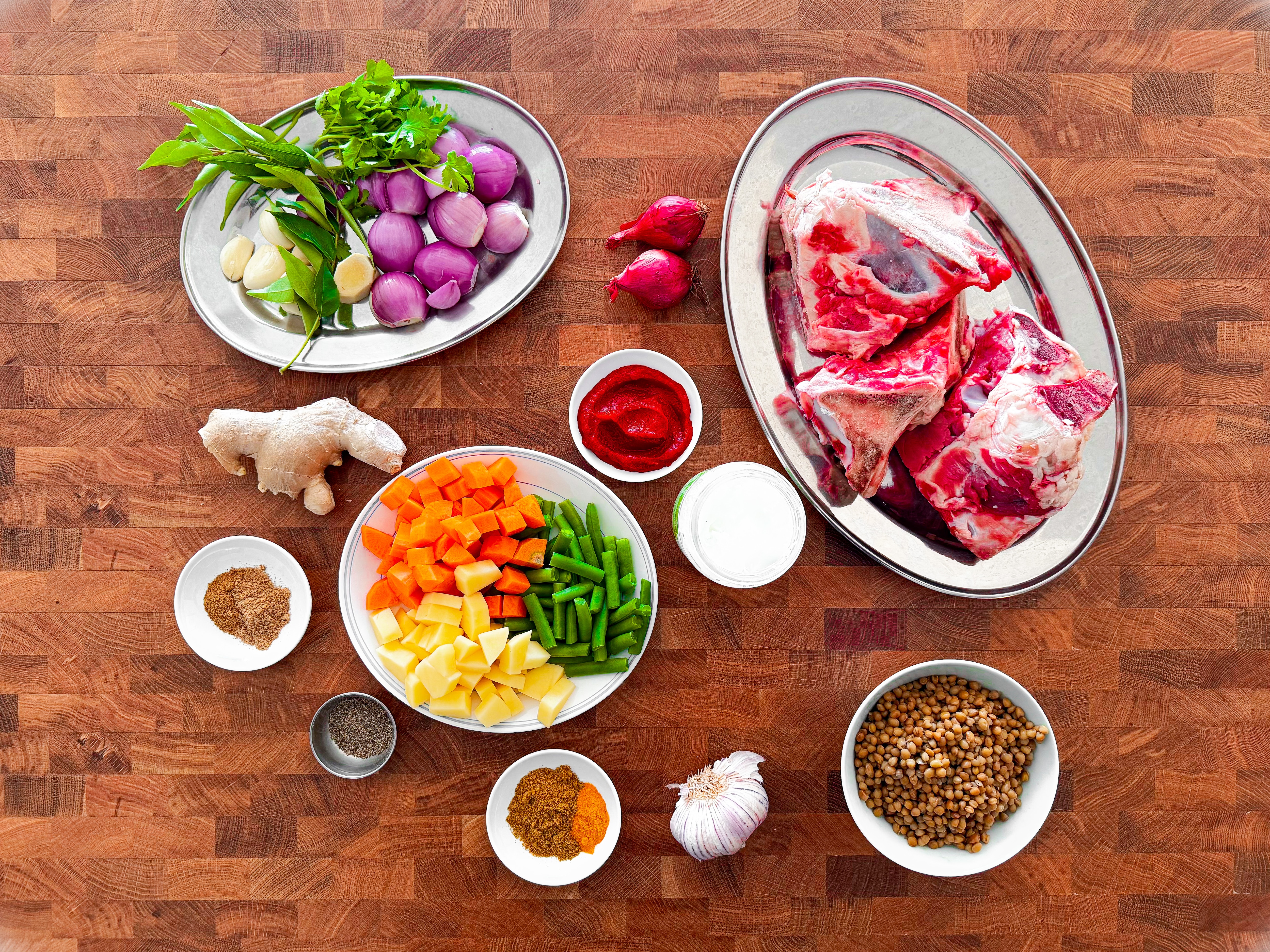 Bowls with ingredients, including beef bones, vegetables, spices and lentils, on a wooden bench.