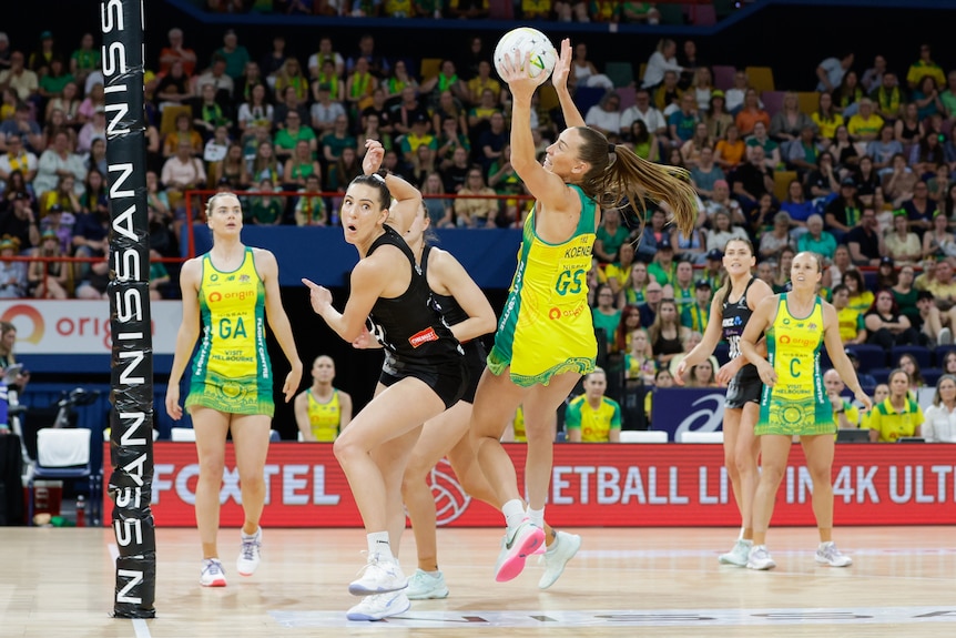 An Australian netballer gets ready to send the ball towards the net as New Zealand defenders look on.