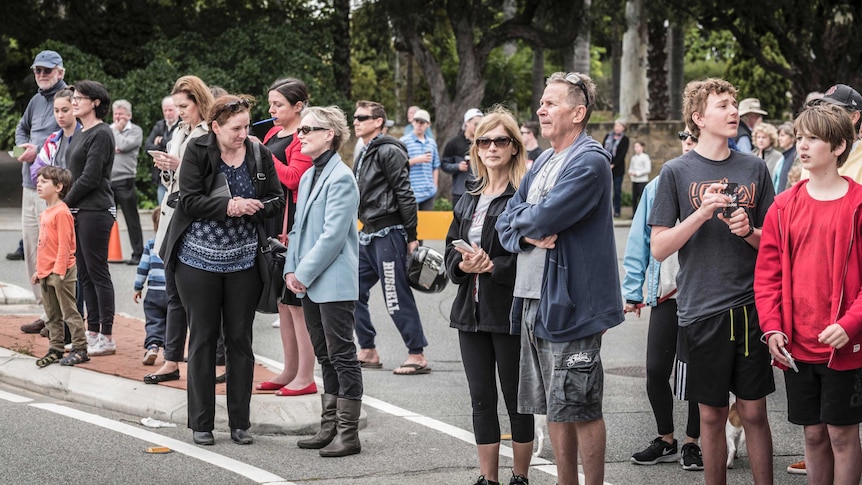 A group of people stand on a road looking twoards an unseen building with cameras and phones.