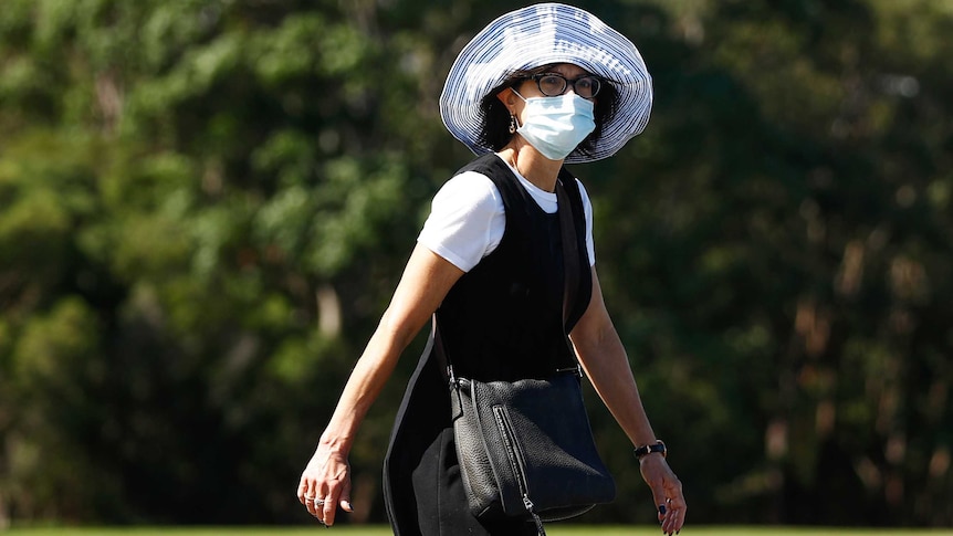 A woman wearing a hat and face mask walks in a park.