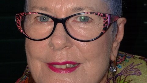 Close-up shot of a woman with short, light purple hair, glasses and a slight smile, with coloured print scarf.
