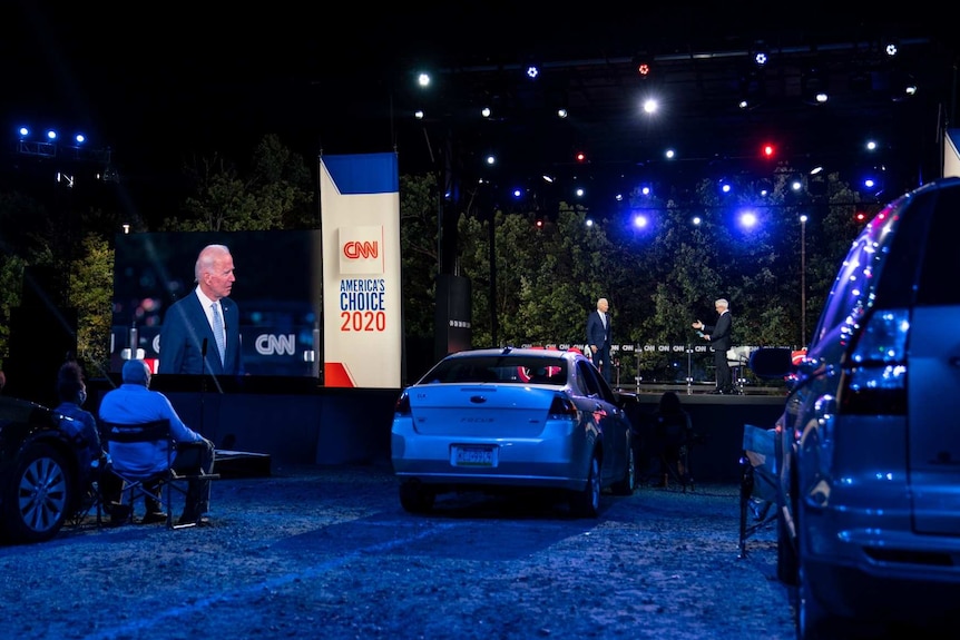 people sit on chairs outside their cars as Joe Biden is shown on stage and on screen at a CNN town hall