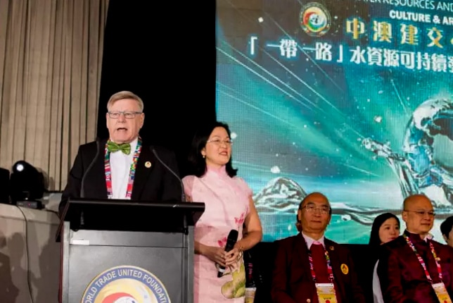 Man wearing suit and bowtie at podium on stage next to women in pink dress, with men and woman dressed formally also on stage