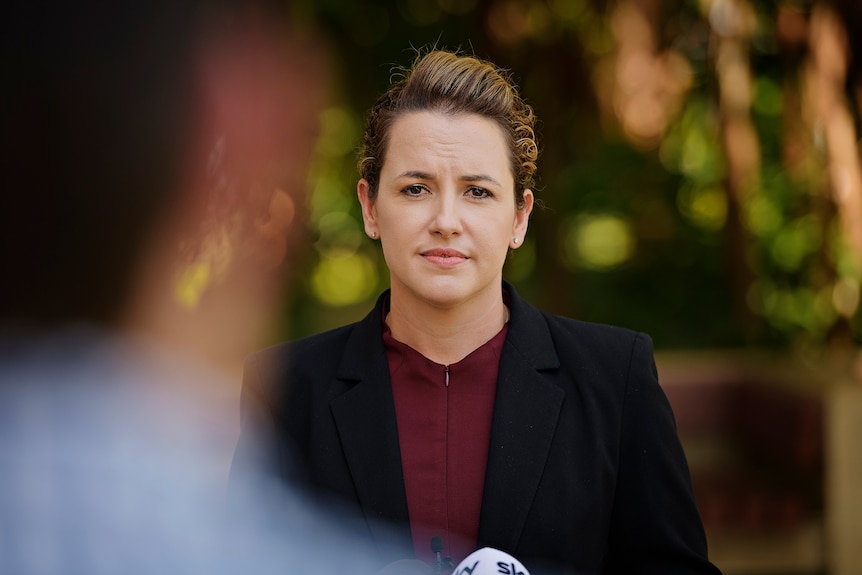 Lia Finocchiaro is wearing a red dress and black blazer and looks seriously at the camera.