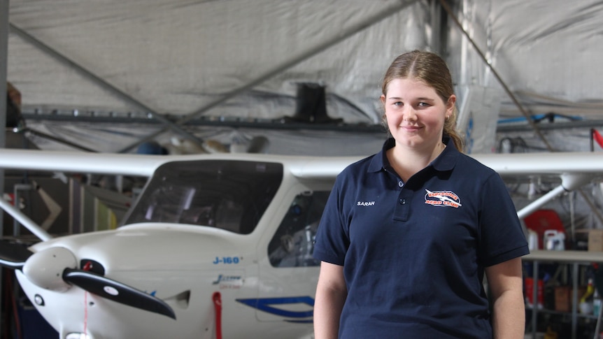 A teenage girl stands in front of a small plane in a hangar. 