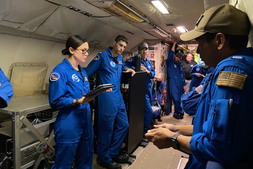A group of people in blue coveralls stand inside a plane looking at a woman who is speaking.