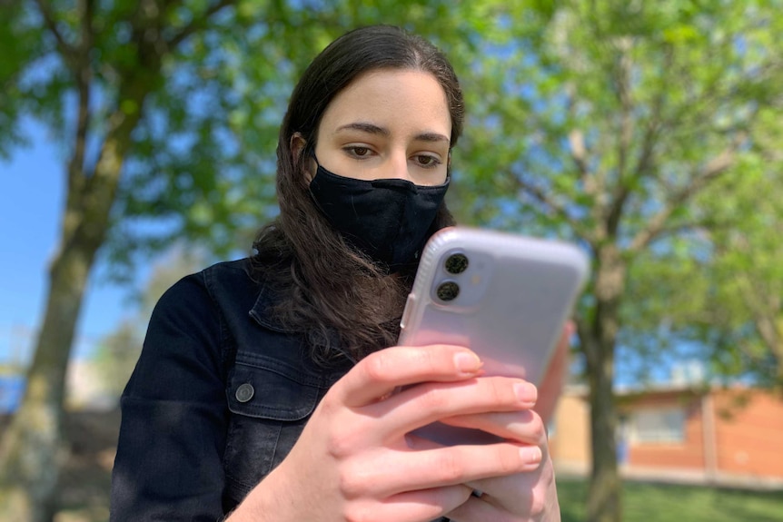 Maya Ghassali sits in a park on her phone, she wears a mask