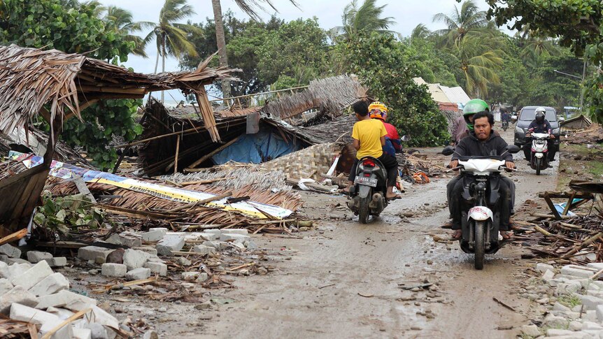 Four men on two scooters are seen driving in opposite directions on dirt road among wreckage left in the wake of a tsunami.