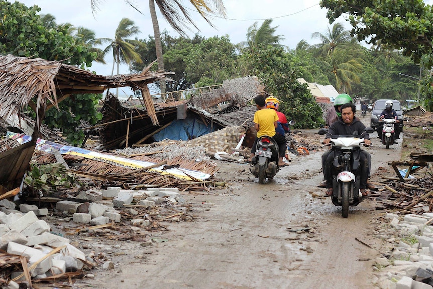 Four men on two scooters are seen driving in opposite directions on dirt road among wreckage left in the wake of a tsunami.