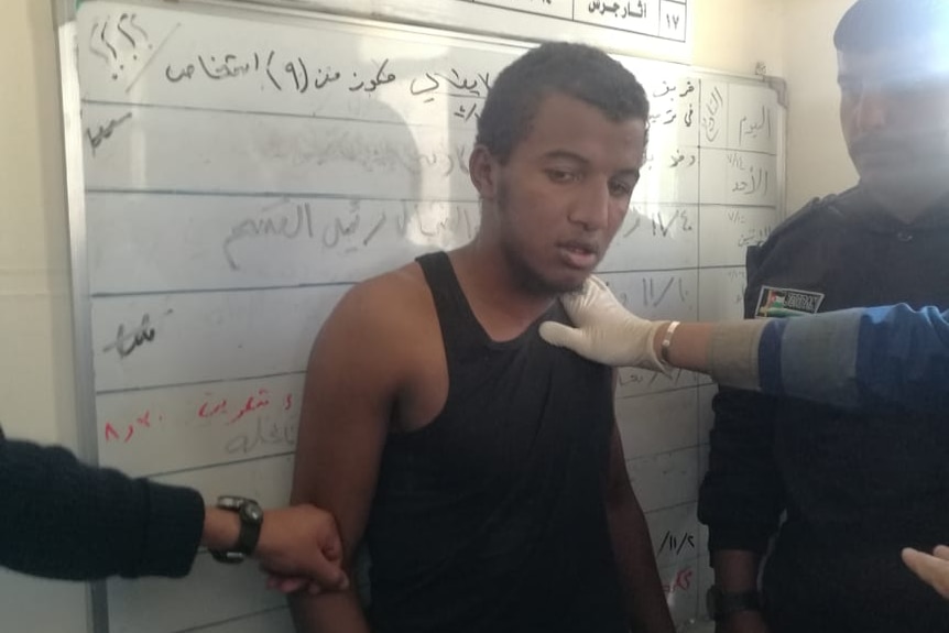 A man is restrained by multiple security guards in a small room as he leans on wall-mounted whiteboards.