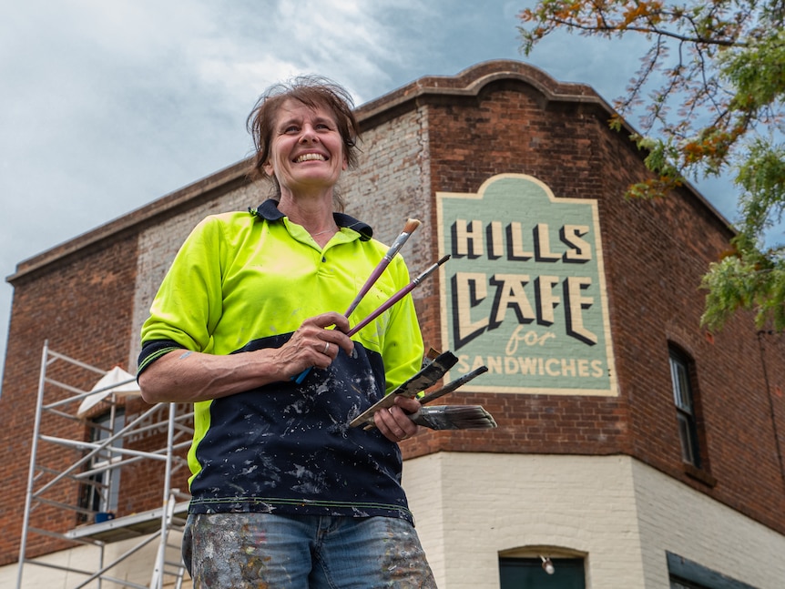 A woman holds paint brushes and smiles in front of a painted sign.