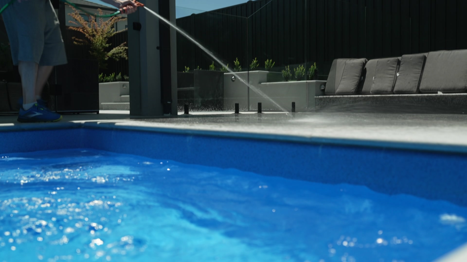 Una persona sostiene la boquilla naranja de una manguera de jardín y rocía agua sobre el concreto junto al agua azul de una piscina.