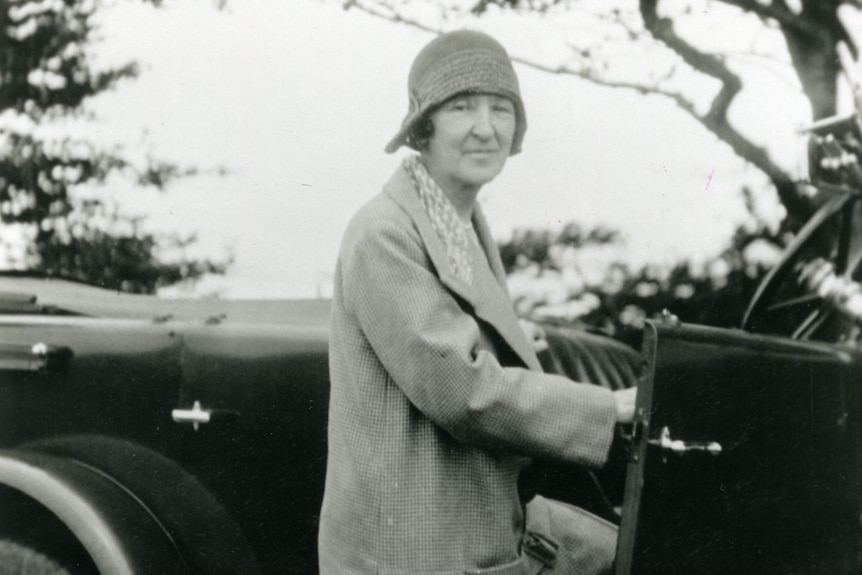 A black and white photo of older woman with a long jacket gets into a old-style car while looking at the camera.