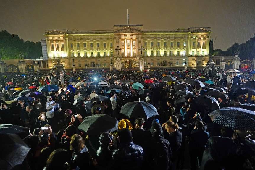 A large crowd gathers outside Buckingham Palace at night,with many holding 