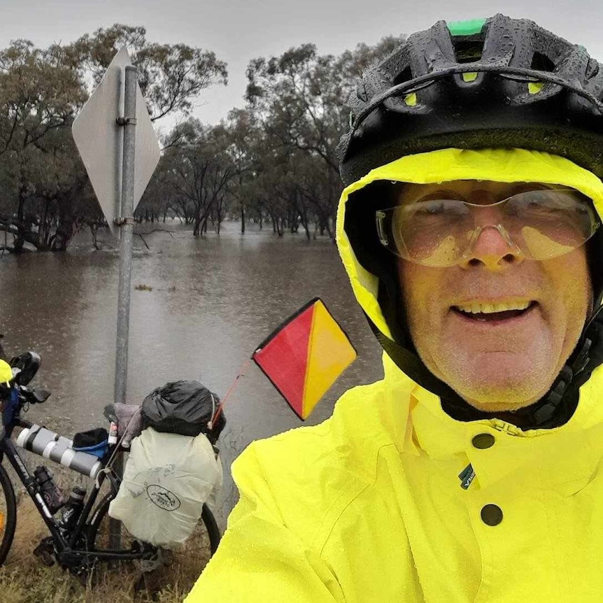 A man in yellow rain coat smiles in front of a pushbike near floodwaters.