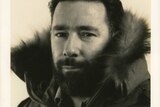 A sepia-toned photo of a man with black hair and a beard looking directly at the viewer, wearing a thick furred-trimmed coat.