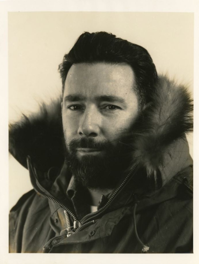 A sepia-toned photo of a man with black hair and a beard looking directly at the viewer, wearing a thick furred-trimmed coat.