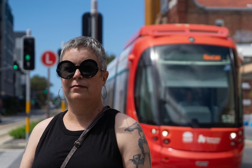 An older woman in all black with short hair and sunglasses stands on a road on a sunny day with a tram in the background