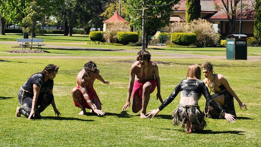 Five First Nations dancers squatting down low outside.