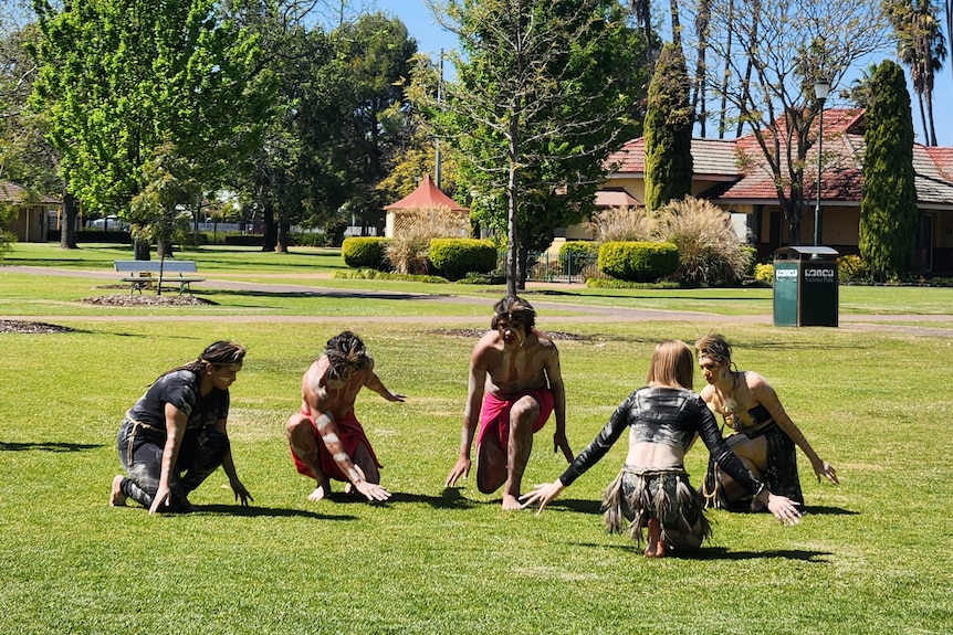 Five First Nations dancers squatting down low outside.