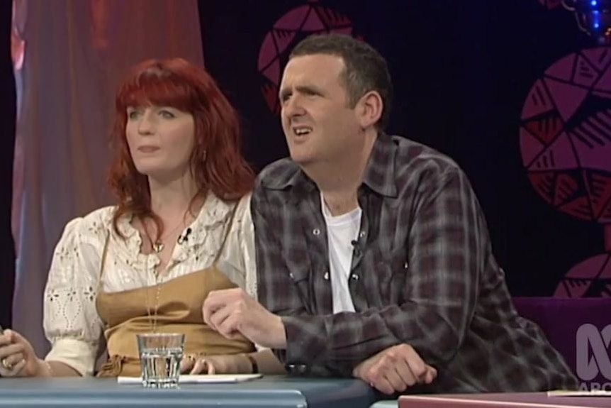 A woman with red hair and a man with brown hair listen while on a panel show on television.