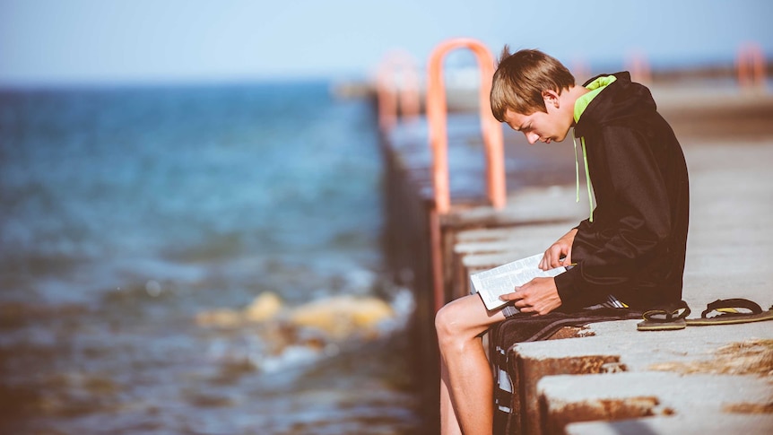 A boy sitting on a jetty reading.