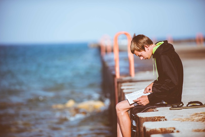 A boy sitting on a jetty reading.
