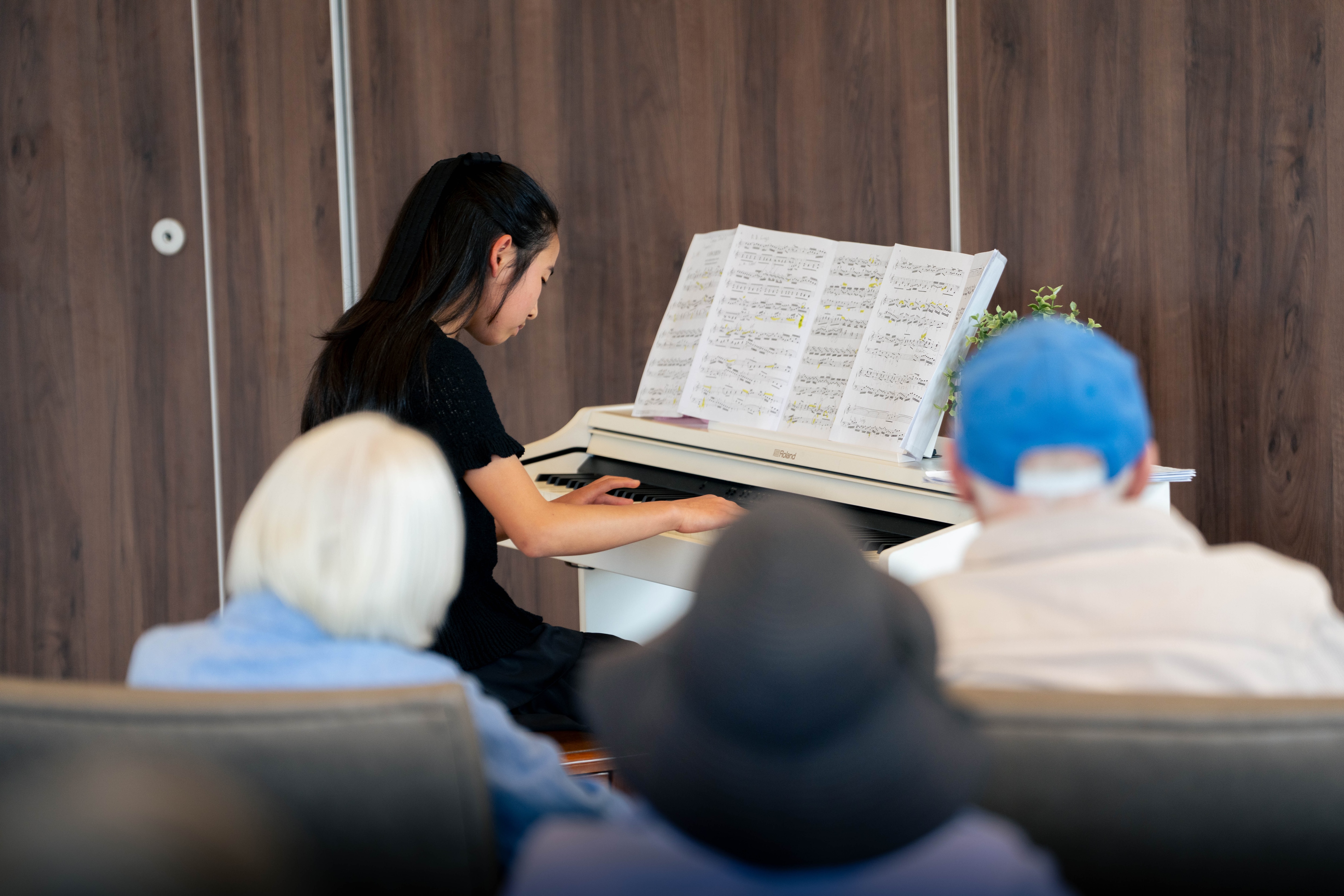 Min, in a black dress, performs a piano piece on a white piano of aged care residents.