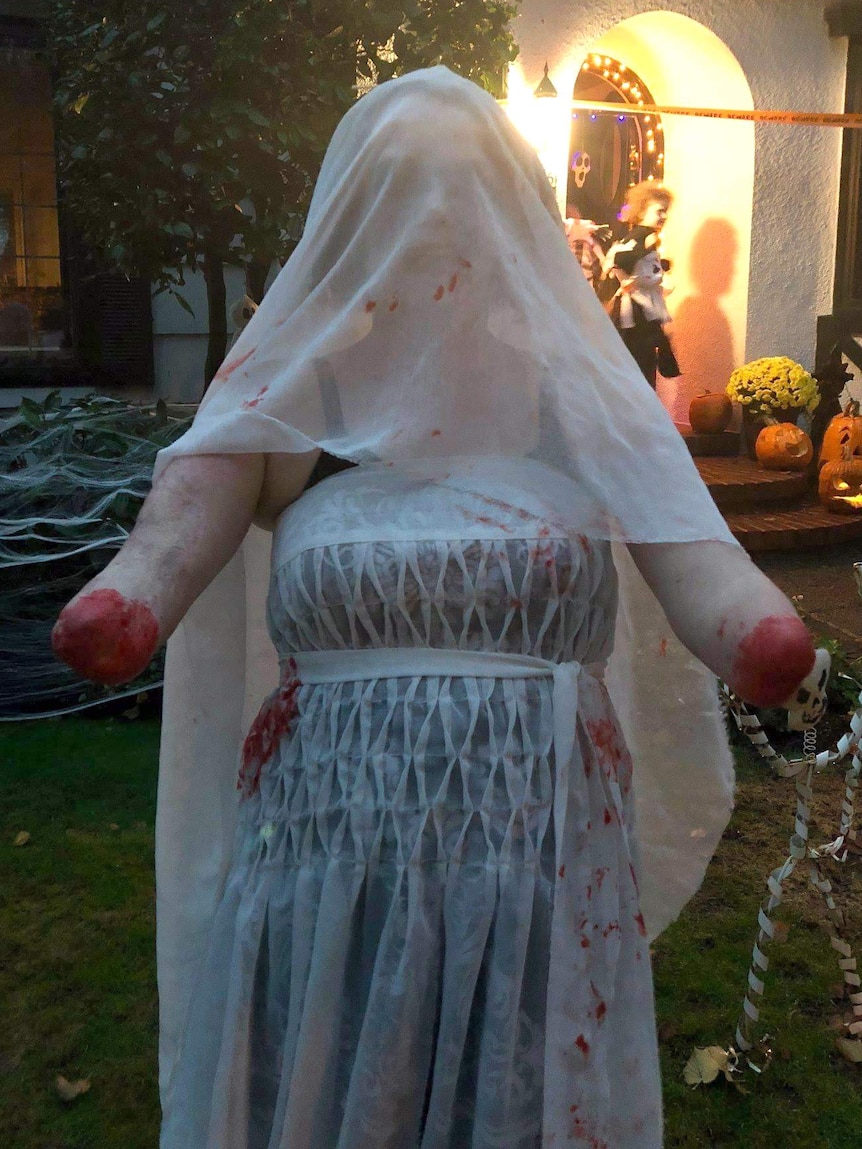 Woman in costume as zombie bride with bloodied dress and veil. She has no hands or forearms. Fake blood painted on ends of arms.