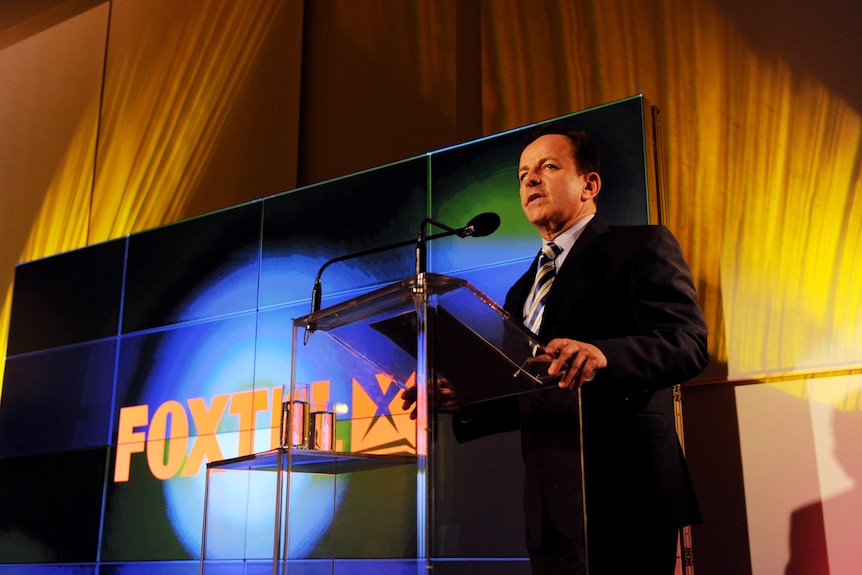 A man in a suit stands behind a lecture with a screen reading "foxtel" behind him