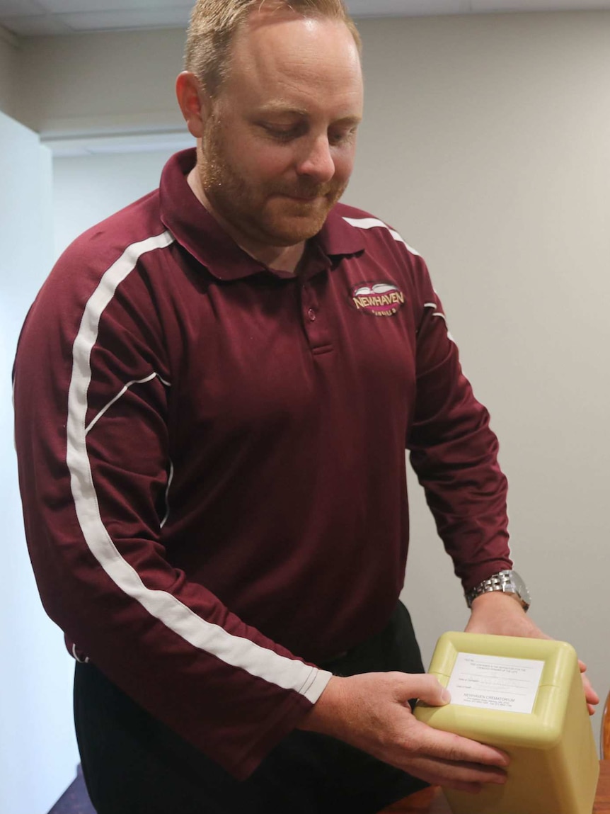 A man with short ginger hair, wearing a maroon tracksuit top with the logo Newhaven, stands holding a box (or urn) on a table.