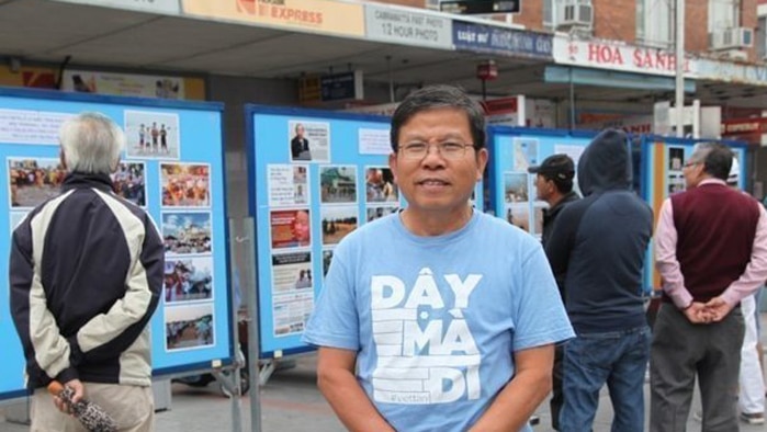 A man of Vietnamese heritage with black hair and glasses stands with his hands clasped in front of of him wearing a blue t-shirt