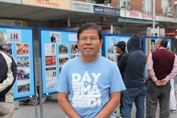 A man of Vietnamese heritage with black hair and glasses stands with his hands clasped in front of of him wearing a blue t-shirt