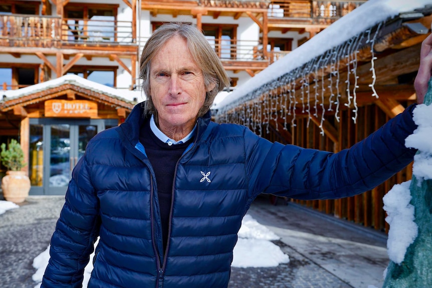 A man in a blue puffer jacket stands outside a picturesque ski chalet