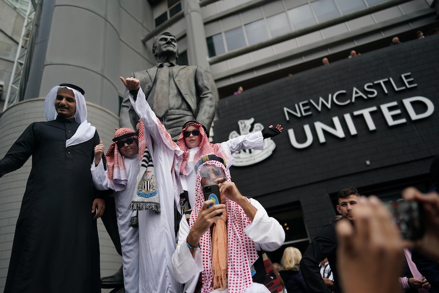 Adults and children wearing Saudi robes and headdresses stand next to the statue of Sir Bobby Robson