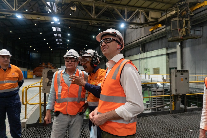 Men in orange vests and hard hats inside a coal plant, pointing.