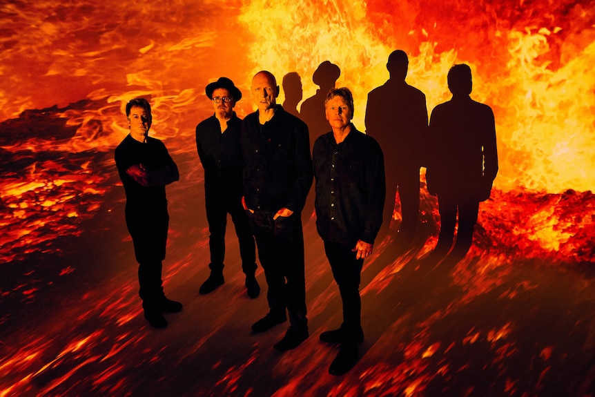 A 2022 press shot of Midnight Oil bathed in firey red light, casting a shadow against the wall