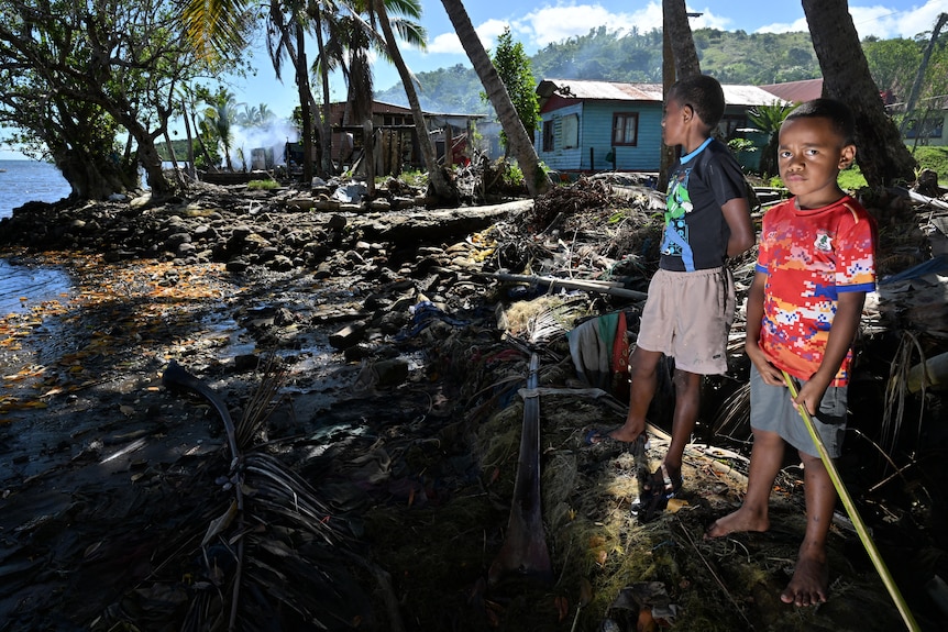 two youths stand on the shore near where their houses stand on the coast of Fiji