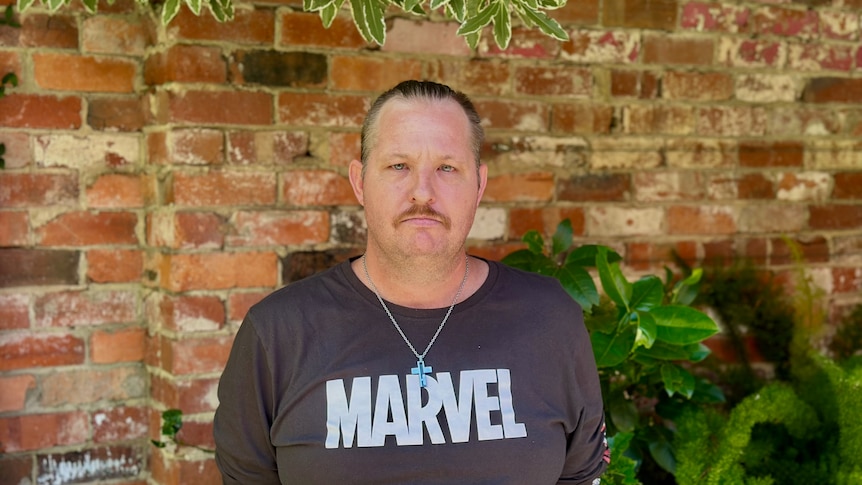 A man wearing a black t-shirt standing in front of a brick wall.