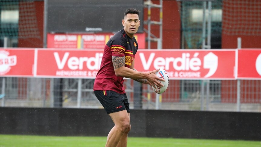 Israel Folau stands in a red and black rugby uniform poised to bass a ball with light rain on the background.