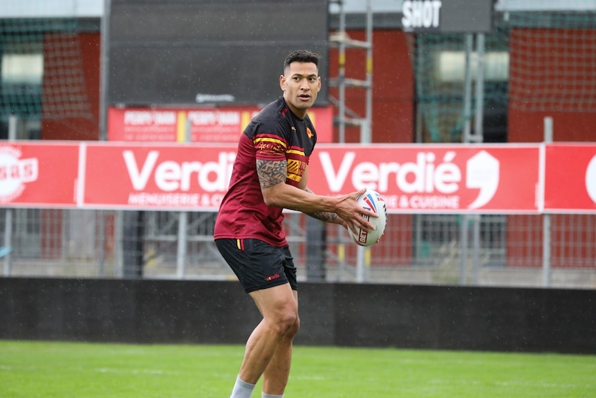 Israel Folau stands in a red and black rugby uniform poised to bass a ball with light rain on the background.