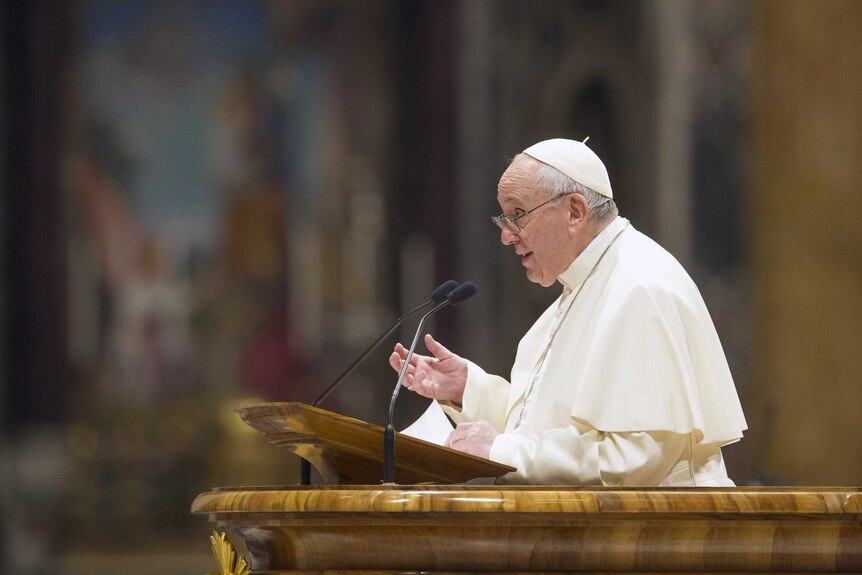 弗朗西斯教皇在木制讲台上身穿白色教皇袈裟的侧视图，背景模糊前有麦克风